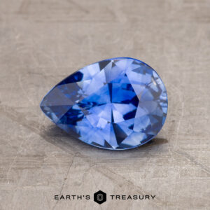 A pear-shaped blue gemstone with multi-faceted cuts is displayed on a textured surface. The text "1.63-Carat Ceylon Sapphire (Heated)" is visible at the bottom of the image.