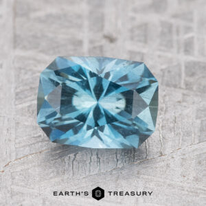 A faceted blue gemstone with a radiant cut placed on a textured background. Text below reads "1.39-Carat Nasarawa Teal Aqua by Earth's Treasury.