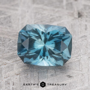 A 1.61-carat Nasarawa Teal Aqua gemstone with many facets, lying on a textured surface. The lower part of the image has the text "EARTH'S TREASURY.