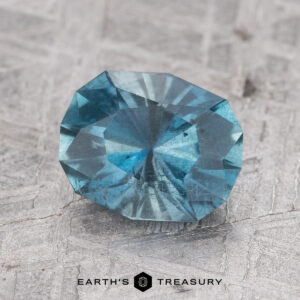 A close-up image of a faceted blue gemstone, the 1.51-Carat Nasarawa Teal Aqua, displayed on a textured gray surface with the text "EARTH'S TREASURY" at the bottom.