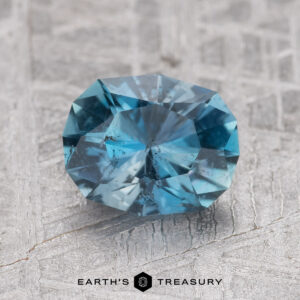 A faceted 1.38-carat Nasarawa Teal Aqua gemstone with slight inclusions is placed on a textured, light gray surface. The text "Earth's Treasury" is visible at the bottom of the image.