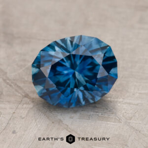 A 2.13-carat faceted blue Montana Sapphire (heated) rests on a textured surface, with the text "Earth's Treasury" appearing at the bottom of the image.