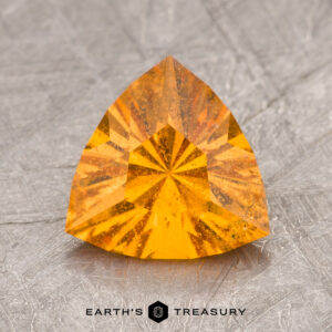 A triangular orange gemstone with a faceted cut, labeled as ET1144, displayed on a textured gray surface.