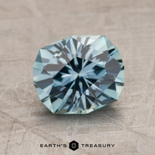 A faceted, round gemstone with a light blue hue rests against a textured, light-colored background. The text "2.18-Carat Montana Sapphire (Heated)" is visible at the bottom of the image.