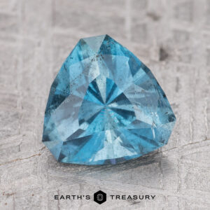 A 1.67-carat Nasarawa Teal Aqua gemstone with a triangular shape sits on a textured surface. The caption "Earth's Treasury" is visible at the bottom of the image.