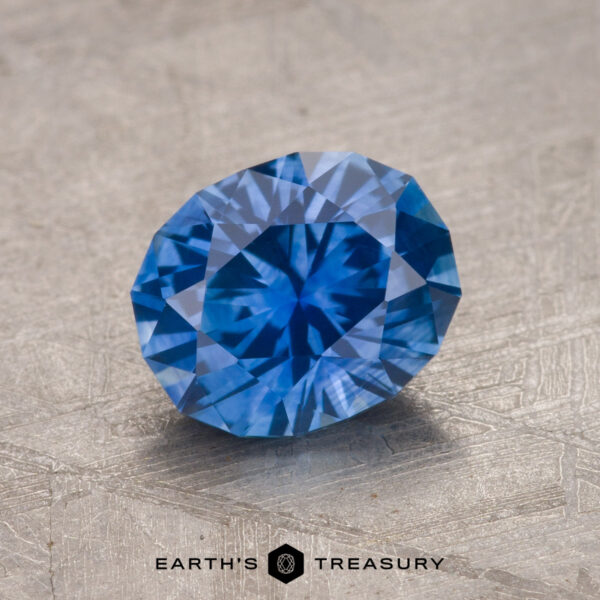 A close-up image of a 1.54-carat rich blue Montana sapphire placed on a textured, metallic surface. The gemstone is expertly cut with multiple facets, reflecting light and showcasing its brilliance. Text at the bottom reads, "EARTH'S TREASURY.