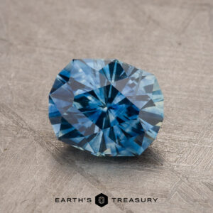 A close-up image of a faceted teal blue Montana sapphire, weighing 1.79 carats and featuring intricate cuts, placed on a textured surface. The bottom center of the image features the text "EARTH'S TREASURY" along with a geometric logo.