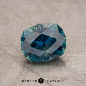 Close-up of a faceted 1.79-carat teal blue Montana sapphire gemstone (heated) placed on a grey, textured surface. The gemstone is cut in a geometric shape, reflecting light with intricate facets. The text at the bottom reads "EARTH'S TREASURY" with a hexagon logo in between the words.