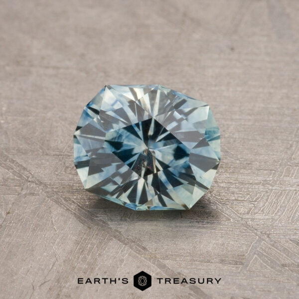 A faceted, round teal blue Montana sapphire weighing 1.79 carats is displayed on a textured gray surface. The gemstone is positioned at an angle, showcasing its intricate cuts and exceptional clarity. The text at the bottom reads "Earth's Treasury.