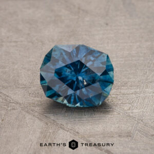 A close-up of a faceted 1.79-carat teal blue Montana Sapphire (heated) gemstone on a textured grey surface. The multifaceted cut enhances the stone's brilliance and depth of color. The logo and text "EARTH'S TREASURY" are seen at the bottom of the image.