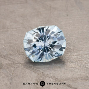 A 1.79-carat teal blue Montana sapphire (heated) is displayed against a textured gray background. The faceted gemstone, with its radiant cut, features intricate facets that catch and reflect light beautifully. The text "EARTH'S TREASURY" is written at the bottom in black.