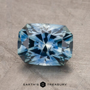 A 1.79-carat teal blue Montana sapphire, heated to enhance its vivid color, rests on a textured surface. The gem boasts intricate faceting that catches the light, creating a sparkling effect. The base of the image displays the text "Earth's Treasury.