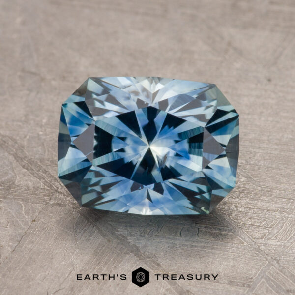 A 1.79-carat teal blue Montana sapphire, heated to enhance its vivid color, rests on a textured surface. The gem boasts intricate faceting that catches the light, creating a sparkling effect. The base of the image displays the text "Earth's Treasury.