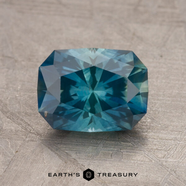 A 1.79-carat teal blue Montana sapphire, featuring a rectangular cut with facets that reflect various shades of blue, is set against a textured beige background. At the bottom, the text reads "EARTH'S TREASURY" alongside a small geometric logo.