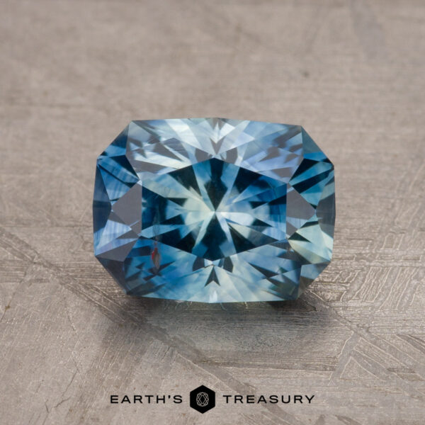 A stunning 1.79-carat teal blue Montana sapphire with a radiant star-like pattern is showcased on a textured surface against a neutral background. The text at the bottom reads "EARTH'S TREASURY".