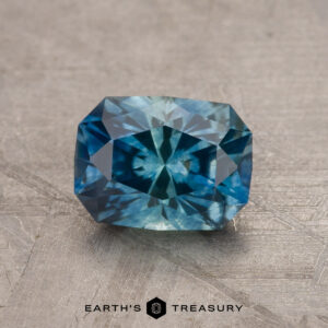 A vibrant 1.79-carat teal blue Montana sapphire (heated) with a rectangular shape and intricate internal facets is displayed on a textured gray surface. The gemstone exhibits a blend of deep and light blue hues, and "Earth's Treasury" is written at the bottom of the image.