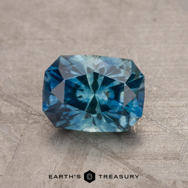 A vibrant 1.79-carat teal blue Montana sapphire (heated) with a rectangular shape and intricate internal facets is displayed on a textured gray surface. The gemstone exhibits a blend of deep and light blue hues, and "Earth's Treasury" is written at the bottom of the image.