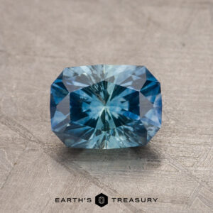 A dazzling, rectangular cushion-cut 1.79-carat Teal Blue Montana Sapphire (Heated) with intricate facets, displayed on a textured gray surface. The gem showcases a vibrant teal blue color with a radiant internal sparkle. Below the gem, the text "EARTH'S TREASURY" is visible.