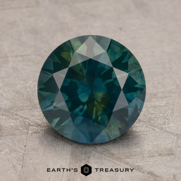 A round, faceted blue-green 2.60-carat Montana Sapphire (Heated) set against a textured gray background, branded Earth's Treasury at the bottom.