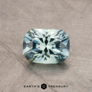 A sparkling rectangular cushion-cut teal blue Montana sapphire gemstone with a 1.79-carat weight is placed on a textured surface. The gem displays high brilliance and intricate facets. The text "Earth's Treasury" is visible beneath the gemstone.