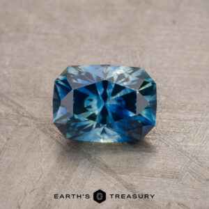 A 1.79-carat teal blue Montana sapphire, featuring a radiant cut style, is displayed against a textured background. The gemstone showcases deep blue and green hues with geometric facets. The text "EARTH'S TREASURY" is visible along the bottom edge of the image.