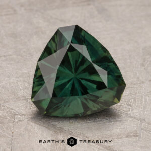 A faceted, dark green 2.37-Carat Australian Sapphire in a triangular cut is displayed on a textured surface with the logo and text "Earth's Treasury" visible below it.