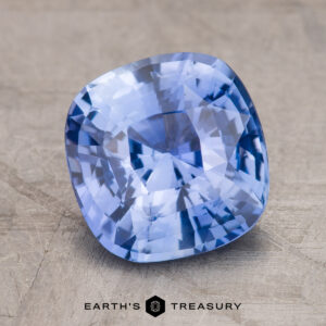 A close-up image of a faceted blue gemstone, expertly captured on a textured background, featuring the words "Earth's Treasury" at the bottom. This stunning gem is the 3.30-Carat Ceylon Sapphire.