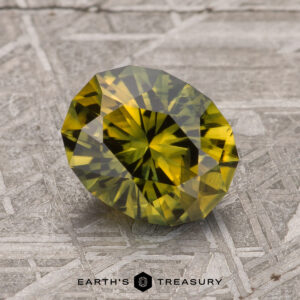 A 2.03-carat green-gold particolored Australian sapphire displayed on a textured, metallic surface. The text "Earth's Treasury" is visible at the bottom of the image.