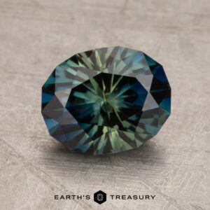 A 2.53-carat Australian sapphire with a faceted oval shape and a blend of green and blue hues is placed on a textured surface. The words "Earth's Treasury" appear below the gemstone.