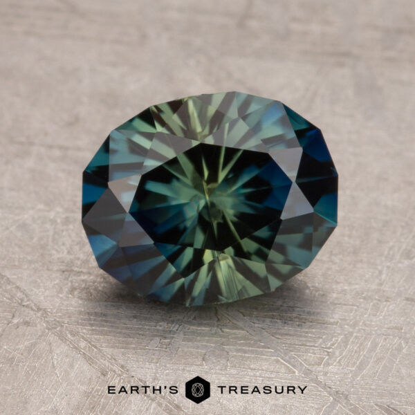 A 2.53-carat Australian sapphire with a faceted oval shape and a blend of green and blue hues is placed on a textured surface. The words "Earth's Treasury" appear below the gemstone.