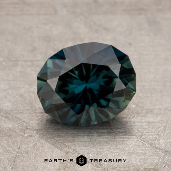 A 1.92-carat Australian sapphire with intricate cuts and a stunning blue-green hue, showcased on a textured gray surface. The gem beautifully reflects light, with the "Earth's Treasury" logo visible at the bottom.