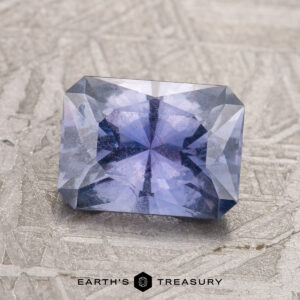 A 2.14-carat violet Umba sapphire, cut in a rectangular shape, sits on a textured surface. The gemstone features a symmetrical internal pattern and is labeled "Earth's Treasury" at the bottom.