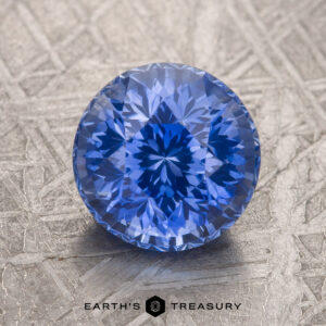 A round-cut blue gemstone is displayed on a textured surface, labeled "3.06-Carat Cornflower Blue Ceylon Sapphire (Heated)" at the bottom.