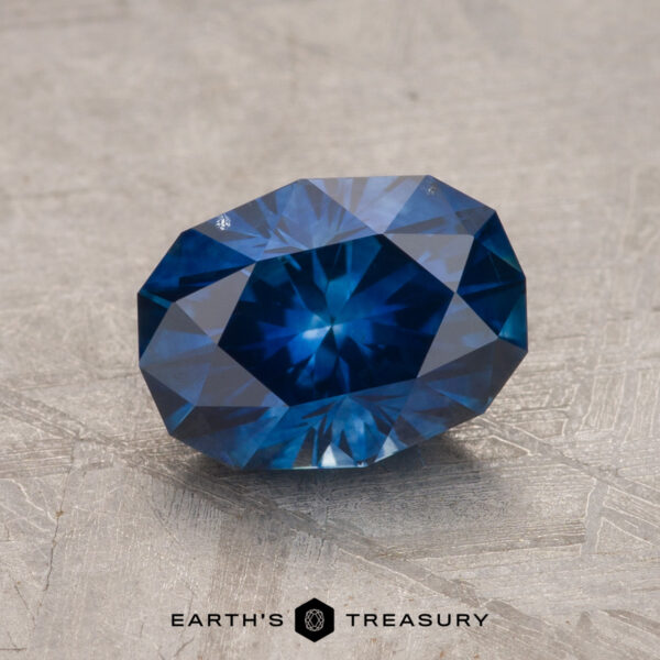 A 2.11-carat Montana sapphire (heated) in deep blue, faceted to perfection, is displayed on a textured grey surface. Below the gem, the text reads "EARTH'S TREASURY.