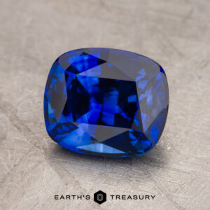 A close-up view of a cut and polished blue 2.54-carat Ceylon Sapphire gemstone on a textured gray surface, labeled "Earth's Treasury" at the bottom.