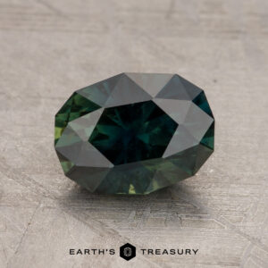 A 2.11-Carat Australian Sapphire, faceted and oval-shaped in dark green, rests on a textured gray surface. Text below the gemstone reads "Earth's Treasury.