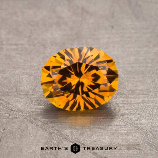 A round, faceted, 1.05-carat Orange Montana Sapphire (heated) with a vibrant, fiery color placed on a textured surface. The background is a grayish metallic material. Text at the bottom reads "Earth's Treasury" with a logo.