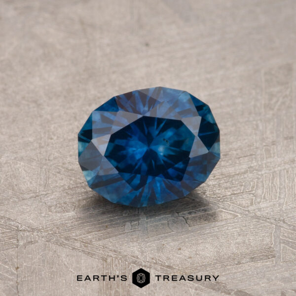 A photo of a 1.01-carat deep blue Montana sapphire (heated) with a rich, captivating color. The gemstone rests on a textured, light-colored surface. The text "EARTH'S TREASURY" appears at the bottom of the image.