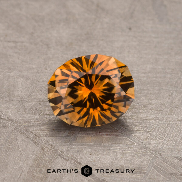 A shimmering round-cut 0.88-carat Orange Montana Sapphire (Heated) with intricate facets, placed on a textured gray surface. The text "Earth's Treasury" is visible at the bottom center of the image.