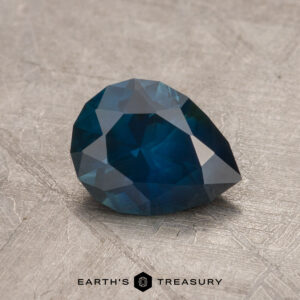 A 1.29-carat faceted Montana Sapphire in a pear shape, displayed on a textured surface, with the text "EARTH'S TREASURY" at the bottom.