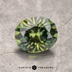 A stunning 2.02-carat Australian Sapphire, round-cut with facets that brilliantly reflect light, is displayed on a textured gray surface. The text "Earth's Treasury" is visible at the bottom center.