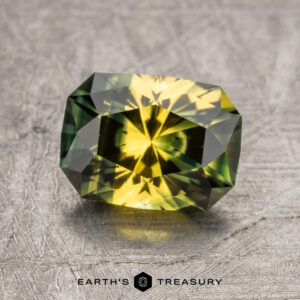 A faceted 1.97-Carat Australian Sapphire gemstone with green and yellow hues, displayed on a textured metallic surface. The text "Earth's Treasury" is visible at the bottom.