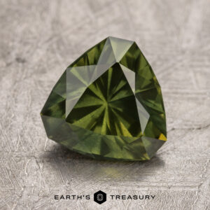 A 2.26-carat Australian Sapphire, cut in a triangular shape with a starburst pattern, is placed on a textured, metallic surface. The words "Earth's Treasury" are visible below the gem.