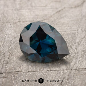 A 1.40-carat Australian sapphire gemstone, featuring a teal color and multiple facets in a pear shape, is placed on a textured surface with the text "EARTH'S TREASURY" visible below it.