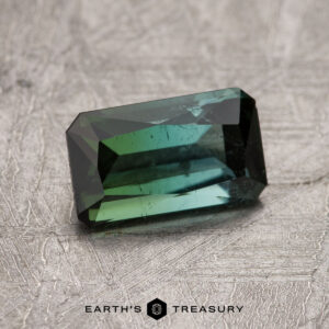 A 1.26-carat California Tourmaline with facets, placed on a textured gray surface. "EARTH'S TREASURY" is written below the stone.