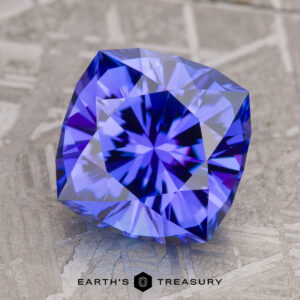 A 3.74-carat rich blue tanzanite, intricately cut with a radiant pattern, lies on a textured surface, displaying bright reflections. The words "Earth's Treasury" are visible at the bottom.