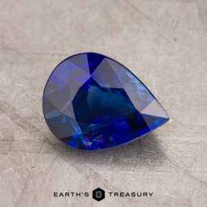 JM1023L features a pear-shaped, blue gemstone with intricate facets set against a textured gray surface, with the text "Earth's Treasury" displayed at the bottom.