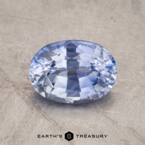 An oval-shaped faceted gemstone with a light blue color, identified as product JM1024L, displayed on a textured surface. The text "EARTH'S TREASURY" is visible at the bottom of the image.