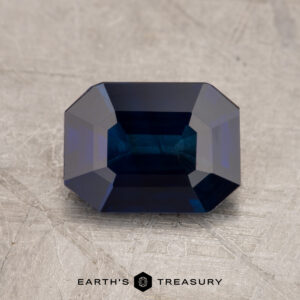 A dark blue, emerald-cut sapphire identified as JM1025L is displayed on a textured gray surface with the "Earth's Treasury" logo below it.