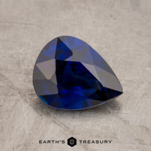 A faceted blue JM1026L gemstone on a textured surface with the "Earth's Treasury" logo at the bottom.